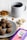 A light grey plate contains several dark brown, round snacks, possibly ragi laddus with visible nut pieces. Nearby, a small bowl of almonds and another with cashew nuts are placed. A blue mug filled with a dark beverage, possibly tea or coffee, is positioned in the background. A purple and pink box labeled 'Bazic Ragi Ladu' is visible in the foreground.