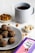 A light grey plate contains several dark brown, round snacks, possibly ragi laddus with visible nut pieces. Nearby, a small bowl of almonds and another with cashew nuts are placed. A blue mug filled with a dark beverage, possibly tea or coffee, is positioned in the background. A purple and pink box labeled 'Bazic Ragi Ladu' is visible in the foreground.