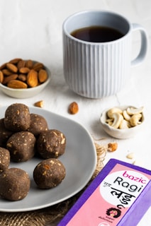A light grey plate contains several dark brown, round snacks, possibly ragi laddus with visible nut pieces. Nearby, a small bowl of almonds and another with cashew nuts are placed. A blue mug filled with a dark beverage, possibly tea or coffee, is positioned in the background. A purple and pink box labeled 'Bazic Ragi Ladu' is visible in the foreground.
