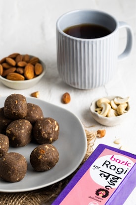 A light grey plate contains several dark brown, round snacks, possibly ragi laddus with visible nut pieces. Nearby, a small bowl of almonds and another with cashew nuts are placed. A blue mug filled with a dark beverage, possibly tea or coffee, is positioned in the background. A purple and pink box labeled 'Bazic Ragi Ladu' is visible in the foreground.