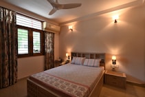 A cozy bedroom with a queen-sized bed featuring a patterned bedspread and two pillows. Soft lighting emanates from wall-mounted lamps on either side of the bed. The room has a wooden look with a bedside table on the right. Curtains with a floral design frame a window, and an air conditioner is mounted above it. The walls are painted in a warm hue, contributing to a serene atmosphere.