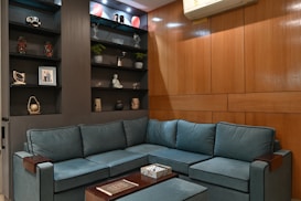 A cozy living room with a teal sectional sofa against a wooden-paneled wall. The adjacent wall features built-in shelves filled with decorative items, including sculptures, plants, and framed art. An air conditioning unit is mounted near the ceiling.