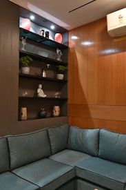 A cozy corner with a teal L-shaped couch against wooden paneled walls. Above the couch is a display shelf featuring various decorative items, including plants in small pots, crystal figurines, a small statue, and artistic vases. The top shelf is illuminated with spotlights, highlighting the objects and two red decorative lamps.