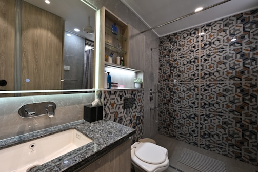 A modern bathroom with geometric patterned tiles on the shower wall, a marble countertop sink, and elegant lighting. Shelves with toiletries and a small plant add a touch of decor, and the overall design features sleek lines and neutral colors.