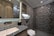 A modern bathroom with geometric patterned tiles on the shower wall, a marble countertop sink, and elegant lighting. Shelves with toiletries and a small plant add a touch of decor, and the overall design features sleek lines and neutral colors.