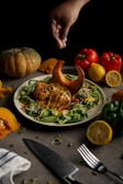 A hand sprinkles seeds over a plate of roasted chicken and salad, featuring a decorative slice of cooked pumpkin on top. The setting includes whole red and green bell peppers, a lemon cut in half, and a whole pumpkin arranged around the plate. A knife and fork are positioned at the edge of the table.