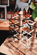 A wooden display stand holds multiple pairs of eyeglasses, with frames in various colors and styles. Behind the stand, several bottles with droppers, likely containing essential oils or tinctures, are arranged on a wooden surface. A large plant with green leaves partially frames the right side of the image, while a paper with text and an image is visible in the background.