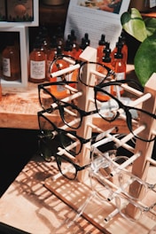 A wooden display stand holds multiple pairs of eyeglasses, with frames in various colors and styles. Behind the stand, several bottles with droppers, likely containing essential oils or tinctures, are arranged on a wooden surface. A large plant with green leaves partially frames the right side of the image, while a paper with text and an image is visible in the background.