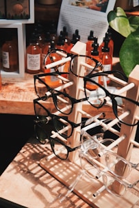 A wooden display stand holds multiple pairs of eyeglasses, with frames in various colors and styles. Behind the stand, several bottles with droppers, likely containing essential oils or tinctures, are arranged on a wooden surface. A large plant with green leaves partially frames the right side of the image, while a paper with text and an image is visible in the background.