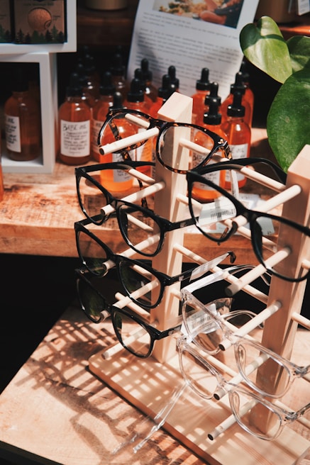 A wooden display stand holds multiple pairs of eyeglasses, with frames in various colors and styles. Behind the stand, several bottles with droppers, likely containing essential oils or tinctures, are arranged on a wooden surface. A large plant with green leaves partially frames the right side of the image, while a paper with text and an image is visible in the background.