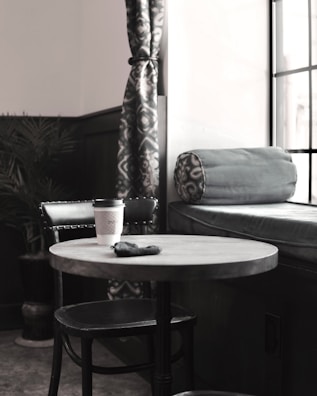 A cozy corner in a cafe with a round table and a single black chair. A coffee cup sits on the table beside a small black pouch. The backdrop features a cushioned bench with a rolled pillow and patterned curtains, complemented by a potted plant.
