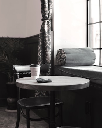 A cozy corner in a cafe with a round table and a single black chair. A coffee cup sits on the table beside a small black pouch. The backdrop features a cushioned bench with a rolled pillow and patterned curtains, complemented by a potted plant.