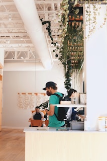 A modern indoor café featuring high ceilings with exposed beams and ductwork. Lush green plants hang from the ceiling, contributing to a natural, serene atmosphere. Several people are present; one wears a cap and backpack while using a smartphone, and two others are seated and appear engaged in conversation or work. The interior design includes light wooden furniture and contemporary artwork on the walls.