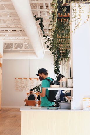 A modern indoor café featuring high ceilings with exposed beams and ductwork. Lush green plants hang from the ceiling, contributing to a natural, serene atmosphere. Several people are present; one wears a cap and backpack while using a smartphone, and two others are seated and appear engaged in conversation or work. The interior design includes light wooden furniture and contemporary artwork on the walls.