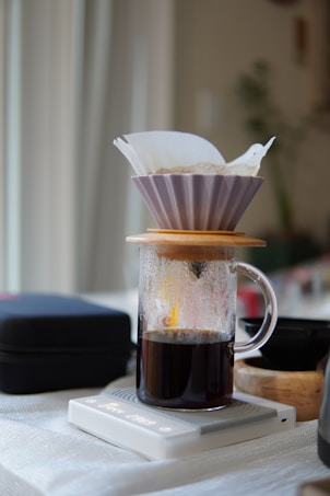 A fresh cup of coffee is being brewed using a drip method. The setup includes a paper filter in a dripper, positioned on top of a glass beaker with coffee collected at the bottom. The setup is placed on a digital scale, against a soft-focused background.