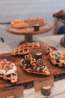 A display of waffles is arranged on a wooden board, each topped with various ingredients such as strawberries, whipped cream, chocolate sauce, pretzels, and chocolate pieces. The presentation is artistic and appetizing, with small label cards in front of the waffles listing their names and prices.