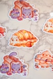 Illustrated stickers featuring whimsical animal-themed pastries and jars labeled 'star jam.' One is a purple cat inside a slice of bread, another is a roll with a white dog, and a third depicts an orange bird with blueberry and crescent moon elements.