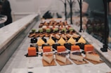 An assortment of colorful pastries and desserts are neatly arranged on a marble countertop. The selection includes pieces of cheesecake, orange and yellow square-shaped pastries, and beautifully decorated eclairs. Bright colors contrast with the soft, muted tones of the counter and background.