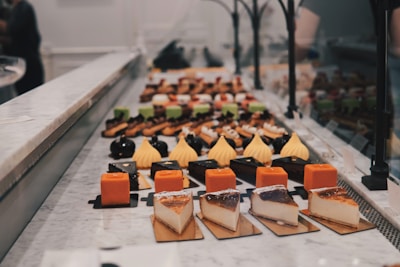 An assortment of colorful pastries and desserts are neatly arranged on a marble countertop. The selection includes pieces of cheesecake, orange and yellow square-shaped pastries, and beautifully decorated eclairs. Bright colors contrast with the soft, muted tones of the counter and background.