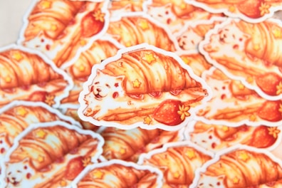 A stack of adorable stickers features a whimsical illustration of a fluffy white dog wrapped in a flaky croissant with strawberries and small star decorations. The artwork combines bright, warm colors and playful elements, creating a cozy and charming aesthetic.