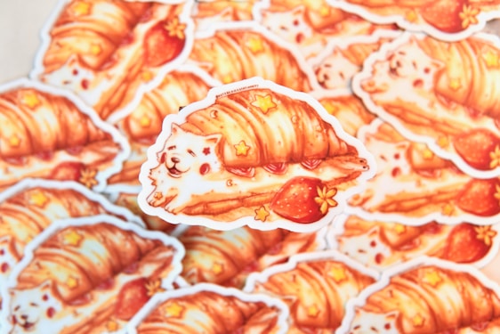 A stack of adorable stickers features a whimsical illustration of a fluffy white dog wrapped in a flaky croissant with strawberries and small star decorations. The artwork combines bright, warm colors and playful elements, creating a cozy and charming aesthetic.