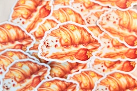 A stack of adorable stickers features a whimsical illustration of a fluffy white dog wrapped in a flaky croissant with strawberries and small star decorations. The artwork combines bright, warm colors and playful elements, creating a cozy and charming aesthetic.