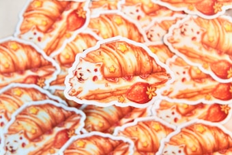 A stack of adorable stickers features a whimsical illustration of a fluffy white dog wrapped in a flaky croissant with strawberries and small star decorations. The artwork combines bright, warm colors and playful elements, creating a cozy and charming aesthetic.