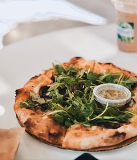 A delicious-looking pizza covered with fresh green arugula is placed on a white table. The pizza has a perfectly browned crust, and a small container of pesto sauce is sitting on top, ready to be added. In the background, there is a to-go cup with a straw, indicating a beverage accompanying the meal.