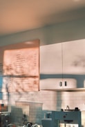 A well-lit café counter area with a wall menu displaying various drink and float options. The light casts soft shadows on the white tile wall and white cabinets. Several kitchen appliances and utensils are visible, providing a cozy and inviting atmosphere.