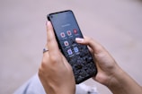 A person is holding a smartphone with their thumbs interacting with the screen. The phone displays a home screen with several social media app icons visible, such as YouTube Studio and Pinterest. The background is a blurred cityscape.