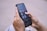 A person is holding a smartphone with their thumbs interacting with the screen. The phone displays a home screen with several social media app icons visible, such as YouTube Studio and Pinterest. The background is a blurred cityscape.