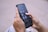 A person is holding a smartphone with their thumbs interacting with the screen. The phone displays a home screen with several social media app icons visible, such as YouTube Studio and Pinterest. The background is a blurred cityscape.