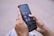 A person is holding a smartphone with their thumbs interacting with the screen. The phone displays a home screen with several social media app icons visible, such as YouTube Studio and Pinterest. The background is a blurred cityscape.