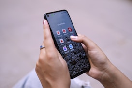 A person is holding a smartphone with their thumbs interacting with the screen. The phone displays a home screen with several social media app icons visible, such as YouTube Studio and Pinterest. The background is a blurred cityscape.