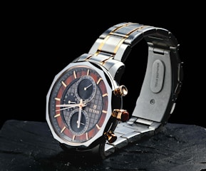 A sleek wristwatch with a stainless steel band featuring gold accents. The watch face combines dark and light elements in a sophisticated design, showcasing several subdials with gold hands and markers. The brand name is visible on the face, adding to its luxurious appeal.