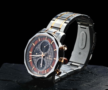 A sleek wristwatch with a stainless steel band featuring gold accents. The watch face combines dark and light elements in a sophisticated design, showcasing several subdials with gold hands and markers. The brand name is visible on the face, adding to its luxurious appeal.