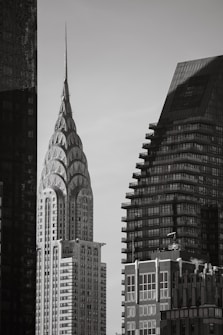 A black and white photograph capturing a section of a city skyline featuring notable skyscrapers. The tallest building has a distinct spire with art deco design elements, while adjacent buildings display a more modern architectural style. The contrast between the architectural designs highlights an urban landscape with historical and contemporary elements.