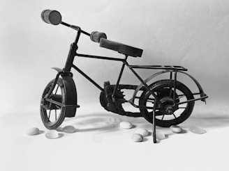 A miniature metal bicycle is set against a plain backdrop. The structure of the bicycle is intricate, with highlights on the frame and gears, creating a vintage appearance. Small white pebbles are scattered around the bicycle, adding contrast to the composition.
