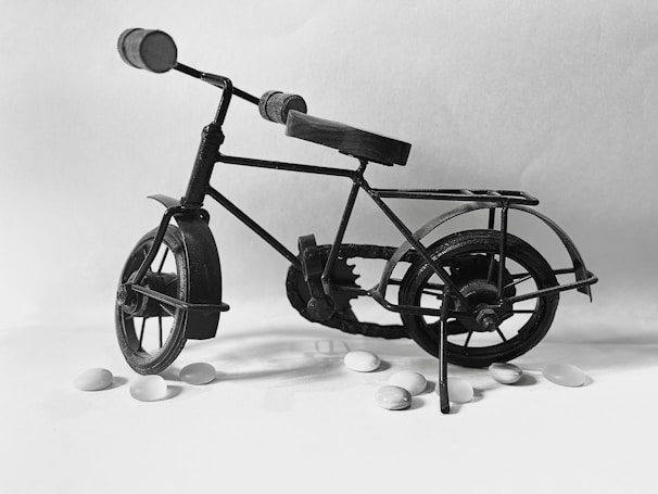 A miniature metal bicycle is set against a plain backdrop. The structure of the bicycle is intricate, with highlights on the frame and gears, creating a vintage appearance. Small white pebbles are scattered around the bicycle, adding contrast to the composition.