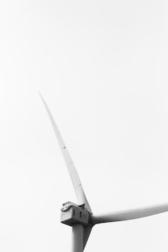 A monochrome image featuring a close-up view of a large, modern wind turbine. The structure is prominently displayed against a clear, featureless sky, emphasizing its sleek and towering design. The turbine's blades are captured in a dynamic angle, showcasing their length and aerodynamic shape.