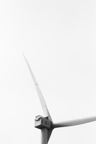 A monochrome image featuring a close-up view of a large, modern wind turbine. The structure is prominently displayed against a clear, featureless sky, emphasizing its sleek and towering design. The turbine's blades are captured in a dynamic angle, showcasing their length and aerodynamic shape.