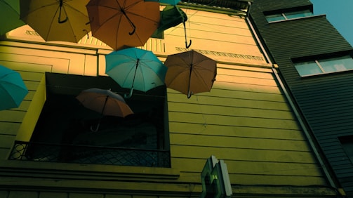 Colorful umbrellas hang upside down against a bright yellow building facade. The umbrellas are suspended on strings and create a playful, whimsical atmosphere. The building has a modern design with large windows and dark trim complementing the vivid colors of the umbrellas.