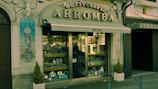A vintage storefront displaying various silverware items behind a glass window. The building facade is ornate with classical elements, including a decorative tile panel featuring a figure holding a clock. The sign above the awning reads 'Ourivesaria Arromba', indicating a jewelry or watch shop. Two small potted plants are placed on either side of the entrance.