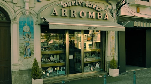 A vintage storefront displaying various silverware items behind a glass window. The building facade is ornate with classical elements, including a decorative tile panel featuring a figure holding a clock. The sign above the awning reads 'Ourivesaria Arromba', indicating a jewelry or watch shop. Two small potted plants are placed on either side of the entrance.