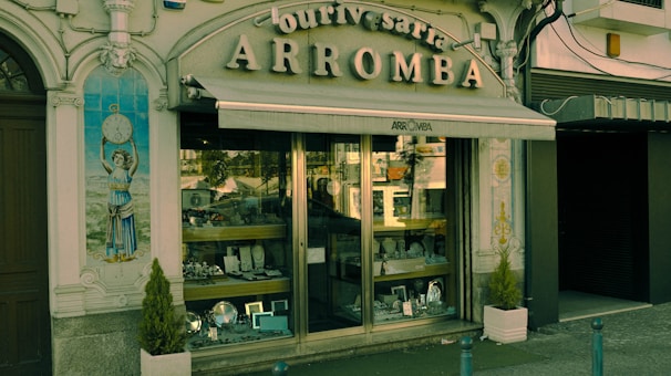 A vintage storefront displaying various silverware items behind a glass window. The building facade is ornate with classical elements, including a decorative tile panel featuring a figure holding a clock. The sign above the awning reads 'Ourivesaria Arromba', indicating a jewelry or watch shop. Two small potted plants are placed on either side of the entrance.