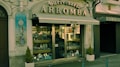 A vintage storefront displaying various silverware items behind a glass window. The building facade is ornate with classical elements, including a decorative tile panel featuring a figure holding a clock. The sign above the awning reads 'Ourivesaria Arromba', indicating a jewelry or watch shop. Two small potted plants are placed on either side of the entrance.
