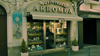 A vintage storefront displaying various silverware items behind a glass window. The building facade is ornate with classical elements, including a decorative tile panel featuring a figure holding a clock. The sign above the awning reads 'Ourivesaria Arromba', indicating a jewelry or watch shop. Two small potted plants are placed on either side of the entrance.