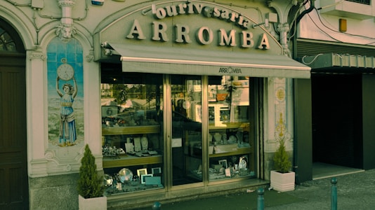 A vintage storefront displaying various silverware items behind a glass window. The building facade is ornate with classical elements, including a decorative tile panel featuring a figure holding a clock. The sign above the awning reads 'Ourivesaria Arromba', indicating a jewelry or watch shop. Two small potted plants are placed on either side of the entrance.