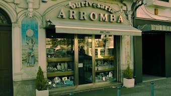 A vintage storefront displaying various silverware items behind a glass window. The building facade is ornate with classical elements, including a decorative tile panel featuring a figure holding a clock. The sign above the awning reads 'Ourivesaria Arromba', indicating a jewelry or watch shop. Two small potted plants are placed on either side of the entrance.