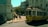 A vintage yellow tram is making its way through a street intersection, surrounded by historic European architecture. Several people are standing near the tram stop; some are taking photos, and others are simply observing. Tall buildings with ornate facades and a few trees can be seen in the background. The lighting suggests it is a sunny day, casting shadows on the ground.